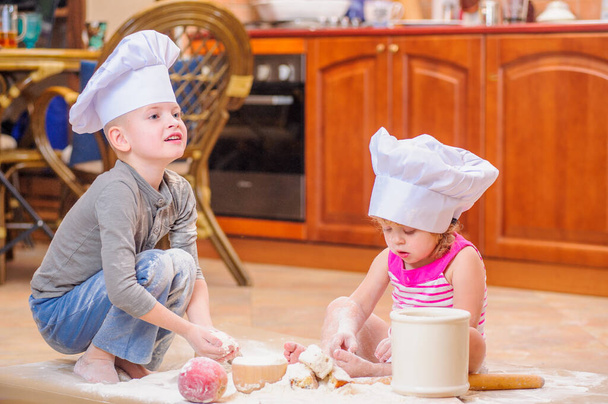 Due fratelli - maschio e femmina - in cappelli da chef vicino al camino seduti sul pavimento della cucina sporchi di farina, che giocano con il cibo, fanno casino e si divertono - Foto, immagini