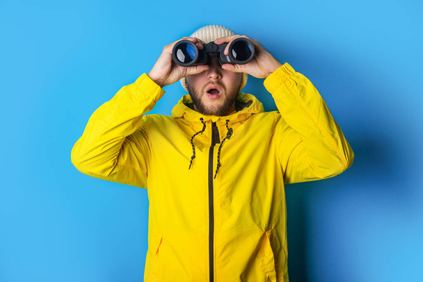 surprised young man in a yellow jacket looks through binoculars on a blue background. - Valokuva, kuva