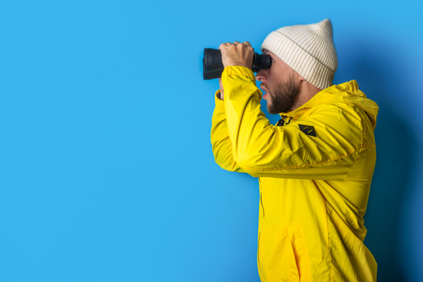 young man in a yellow jacket looks through binoculars on a blue background. - Valokuva, kuva