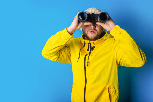 young man in a yellow jacket looks through binoculars on a blue background. - Valokuva, kuva