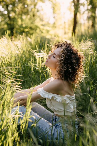 Jeune femme aux cheveux bouclés courts, assise sur de l'herbe verte dans un parc, relaxante. - Photo, image