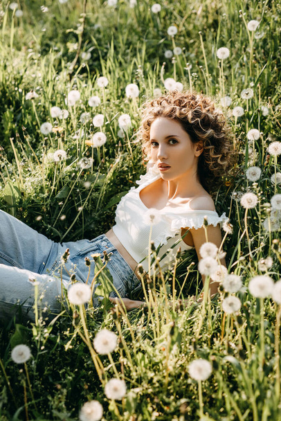 緑の芝生とタンポポの牧草地に横たわっている巻き毛の若い女性. - 写真・画像