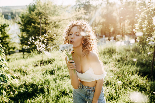 junge Frau mit lockigem Haar pustet an einem Sommertag Löwenzahn in einem Park. - Foto, Bild