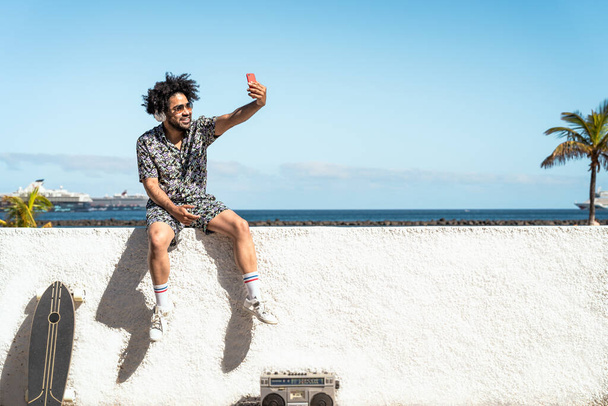 Jeune homme latin s'amuser à prendre selfie avec smartphone mobile tout en écoutant de la musique avec écouteurs et boombox pendant les vacances d'été - Photo, image
