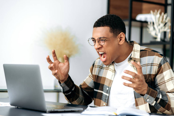 Angry annoyed hispanic young man with glasses, freelancer, manager or student, emotionally yells at a laptop, got bad news, failed, lost, gesturing with hands, very irritated and frustrated - Foto, imagen