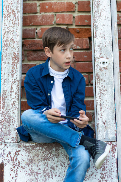Egy tinédzser fiú kék ingben és farmerben okostelefonnal a kezében ül egy régi ajtón, téglafalas háttérrel szemben. Szelektív fókusz - Fotó, kép