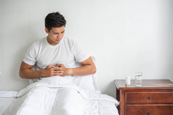 Um homem senta-se na cama com uma dor de estômago e pressiona o estômago com as mãos. - Foto, Imagem