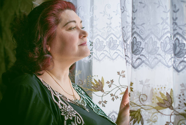 Mujer de mediana edad frente a las cortinas y mirando la vista desde una ventana. Solo más tamaño mujeres maduras, problemas entre las mujeres - Foto, imagen