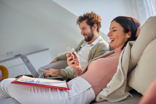 young caucasian couple sitting casually at sofa, smiling, working. Woman looking at her cell phone laughing. - Photo, image
