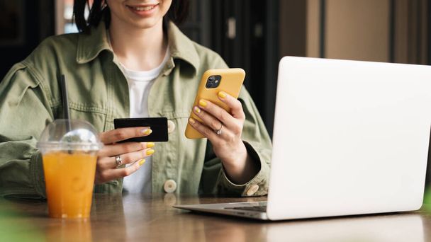 Smiling Unrecognizable Woman Holding Credit Card and Smartphone Making Online Shopping While Sitting at a Cafe Table with Laptop and Cocktail. - Фото, изображение