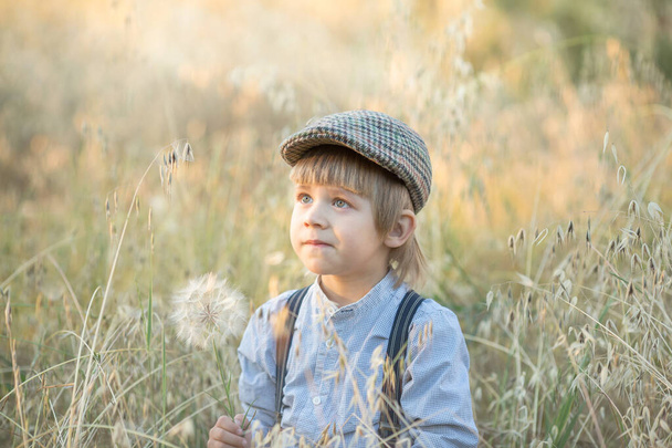 the boy sits in a field and looks up in surprise. in his hands is a dandelion - Photo, Image