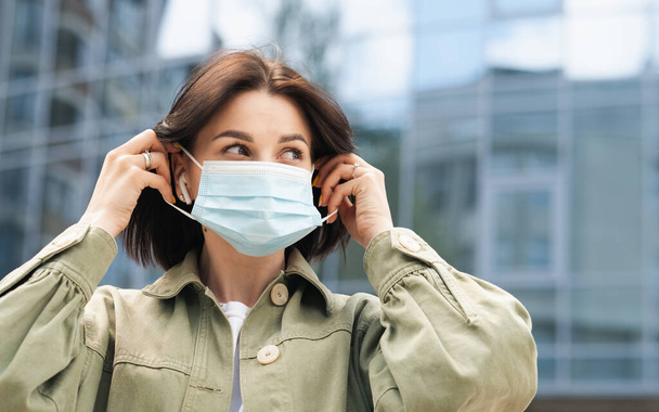 Young Woman Putting a Protective Face Mask on Her Face Because of Pandemic, Portrait of Brunette Girl Using Earphones Standing Outdoors Against City Buildings. - Photo, Image