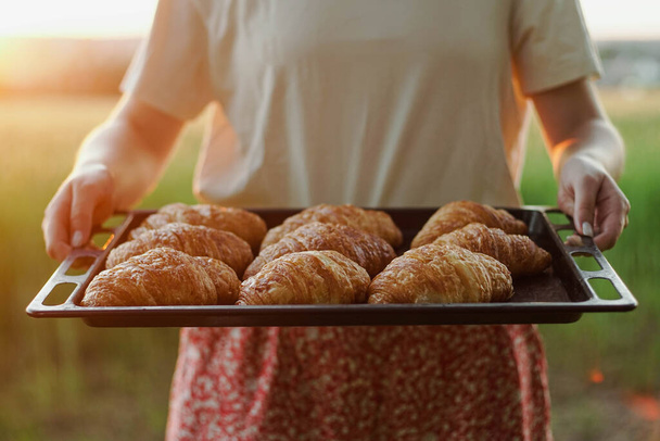 Female baker holding metal tray with fresh croissants at sunset, close up - Photo, Image