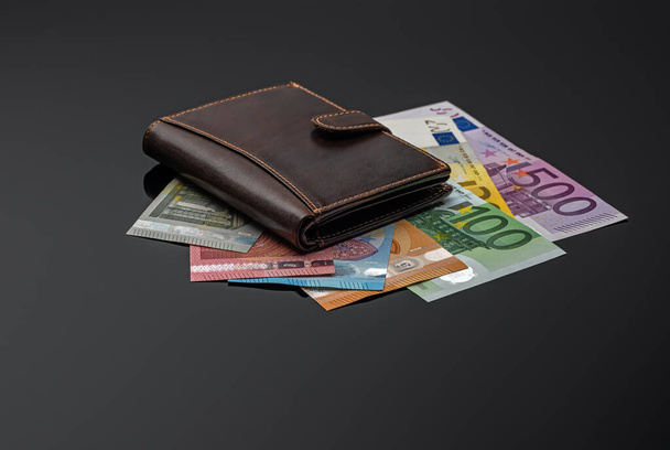 A brown leather wallet rests on banknotes and a reflective gray surface. The main object is highlighted and the reflective surface is shaded with a gradient from left to right. Shallow depth of field. Free place. Low key - Photo, Image