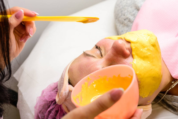 Εφαρμόζοντας μια χρυσή μάσκα στο πρόσωπο μιας γυναίκας, μια διαδικασία σε ένα σαλόνι ομορφιάς. νέα - Φωτογραφία, εικόνα