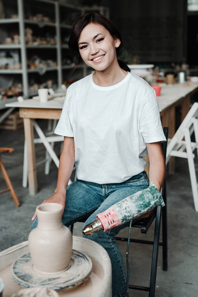 Cute young asian girl in a t-shirt and jeans sits on a chair making a clay vase on a potters whee - Foto, immagini