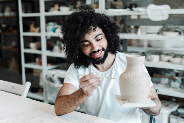 Happy african american man in a t-shirt holds a clay vase in his hand and paints it with a brush - 写真・画像