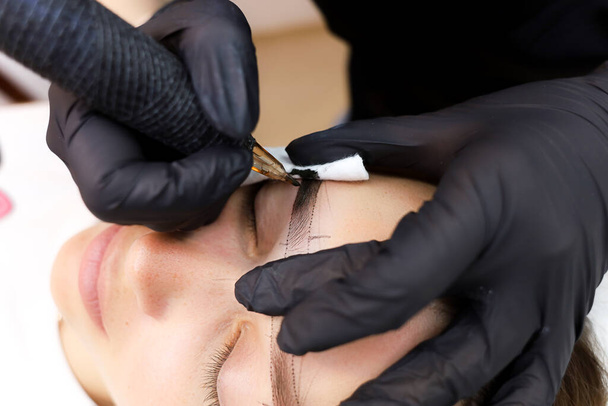 pysyvä meikkitaiteilija suorittaa kulmakarvojen tatuoinnin pitämällä kulmakarvaa kädellään ja venyttämällä ihoa - Valokuva, kuva
