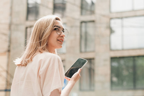 Back Pohled na elegantní ženu v brýlích Držící Smartphone v ruce a usmívající se, stylové blondýny dívka s brýlemi stojící venku na ulici. - Fotografie, Obrázek