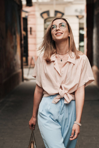 Jonge elegante vrouw in glazen wandelen in de stad, stijlvol blond meisje met bril kruisen op straat buiten, op zoek naar. - Foto, afbeelding