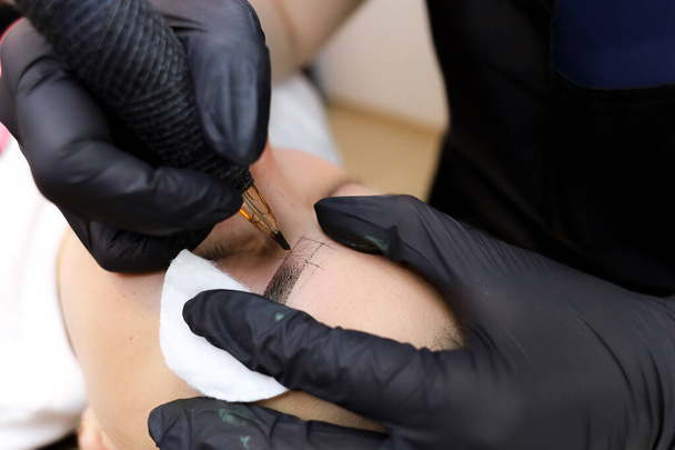 az állandó smink mestere a szemöldökökön tetoválást végez egy tetoválógéppel, amely a szemkörnyéket pamut szivaccsal tartja - Fotó, kép