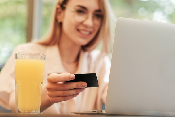Blurred Young Smiling Woman Using Laptop for Online Shopping While Sitting in Restaurant, Blonde Girl with Glasses Holding Credit Card and Making Payment Through the Internet, Εστίαση στην πιστωτική κάρτα. - Φωτογραφία, εικόνα