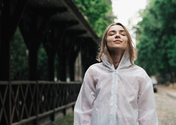Close Up Portrait of Young Woman Wearing Raincoat While Standing on the Street at Rainy Day, Blonde Girl Dressed in Waterproof Cloak Having Fun Under Rain in City. - 写真・画像