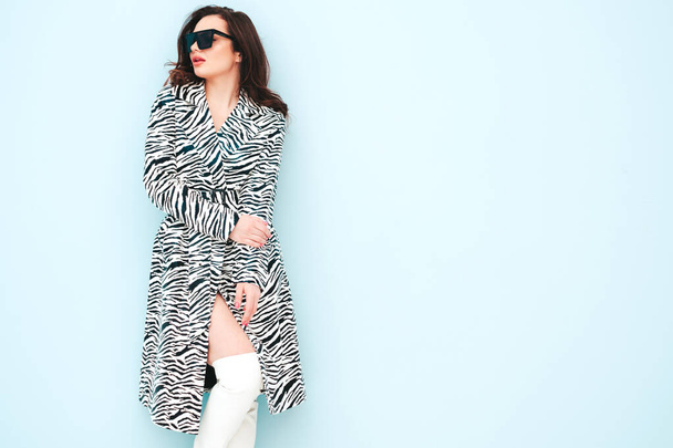 High fashion portrait of young beautiful brunette woman wearing nice trendy summer zebra coat.Sexy fashion model posing near light blue wall in studio.Fashionable female indoors in sunglasses - Photo, image