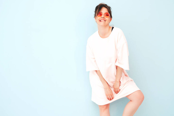 Young beautiful smiling female in trendy summer hipster clothes. Sexy carefree woman posing near light blue wall in studio. Positive model having fun indoors. Cheerful and happy in sunglasses - 写真・画像