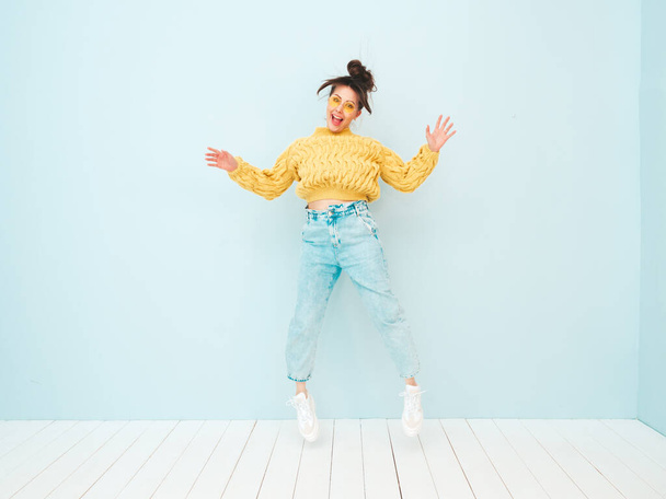 Young beautiful smiling female in trendy summer yellow hipster sweater and  jeans. Sexy carefree woman posing near light blue wall in studio. Positive model having fun indoors. She jumping - Photo, Image