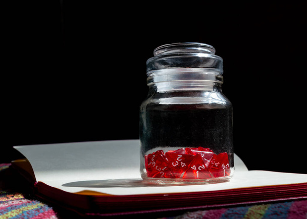 Ein mit roten 4-seitigen Würfeln gefülltes Glas auf einem offenen Notizbuch im Sonnenlicht - Foto, Bild