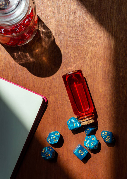 Vertical overhead image of a set of blue marbled role-playing dice, a glass bottle with red liquid, a notebook and, a glass jar filled with red dice - Photo, Image