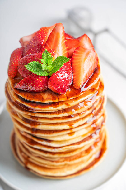 Closeup view of homemade american tasty pancakes or crepes decorated with ripe fresh red strawberries and green mint leaves topping served in stack on gray round plate on white wooden table. Vertical  - Photo, Image