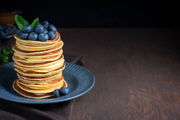 Pile of hot homemade american sweet pancakes or crepes decorated with blueberries and mint leaves served on blue gray plate on dark wooden background with textile. Image with copy space, horizontal - Фото, изображение