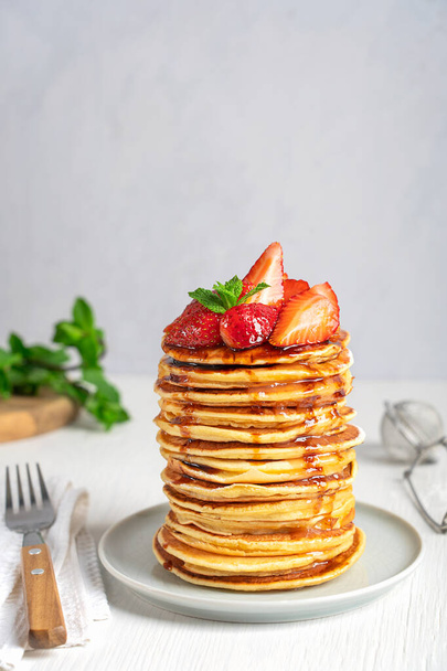 Portion of homemade american organic pancakes or crepes decorated with strawberries and mint leaves served in stack on gray round plate on white wooden table with fork. Vertical image with copy space - Фото, изображение