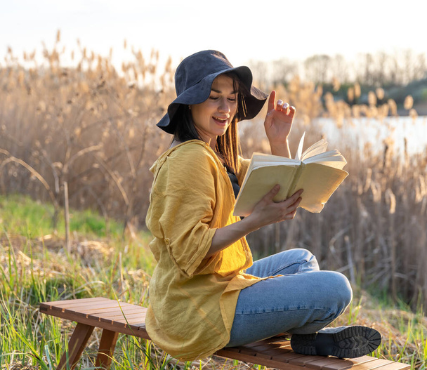 Chica atractiva en un sombrero lee un libro en la naturaleza al atardecer. - Foto, Imagen