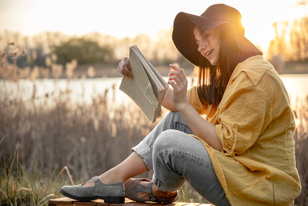 Eine junge Frau mit Hut und einem Lächeln im Gesicht liest bei Sonnenuntergang ein Buch am Fluss.. - Foto, Bild