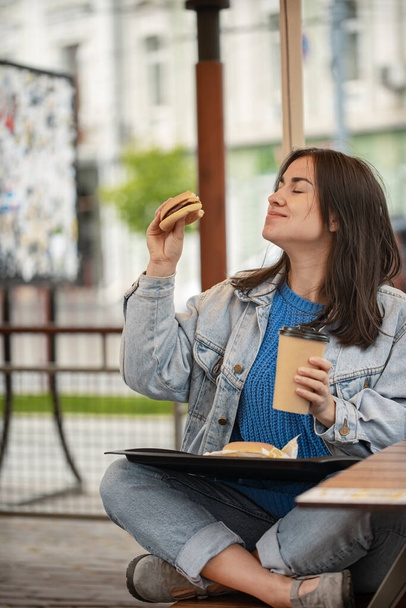 Attraente ragazza in stile casual mangia un hamburger con caffè seduto sulla terrazza estiva. - Foto, immagini