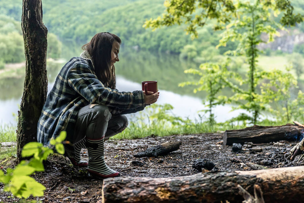 Una giovane donna nella foresta vicino al fiume si scalda vicino al fuoco e beve una bevanda calda. - Foto, immagini