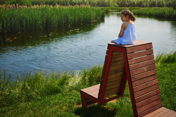 Una linda chica en un vestido azul y dos colas de caballo se sienta en la parte superior de un banco de madera y felizmente observa una hermosa vista de la perspectiva saliente del río en el campo - Foto, Imagen