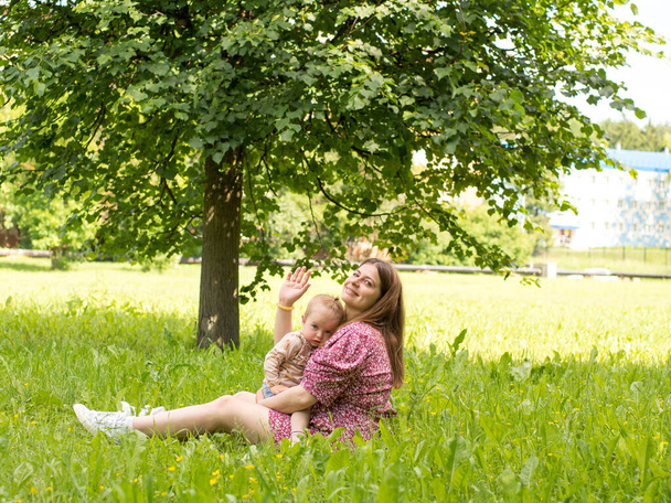 schöne junge Mutter mit einem Baby auf dem Arm sitzt an einem sonnigen Tag im Park auf dem Rasen - Foto, Bild