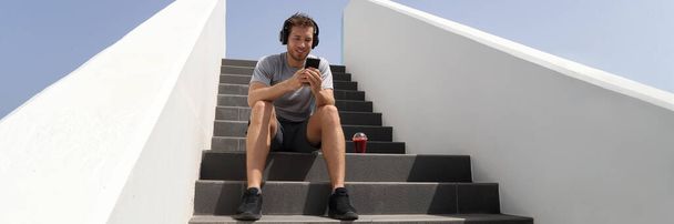 Man listening to music mobile phone app with headphones on stairs banner. Healthy city lifestyle person using smartphone on jogging break with morning red beet juice smoothie sitting outdoors - Foto, afbeelding