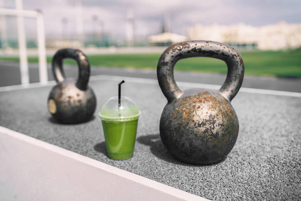 Health and fitness green smoothie detox drink at gym with kettlebells weights at outdoor training fitness center. Plastic cup of vegetable juice morning breakfast next to kettlebell weight equipment - Fotografie, Obrázek