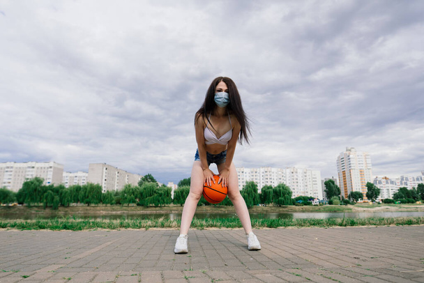 Jovem esporte fitness mulher jogador de basquete usando máscara para proteger com bola, fundo do lago. - Foto, Imagem
