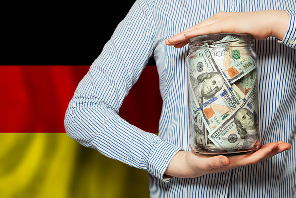 Δολάρια ΗΠΑ μετρητά σε γυάλινο βάζο στο χέρι σε φόντο γερμανική σημαία. Έννοια των επιχειρήσεων, της πολιτικής, των τραπεζών και της διαφθοράς - Φωτογραφία, εικόνα