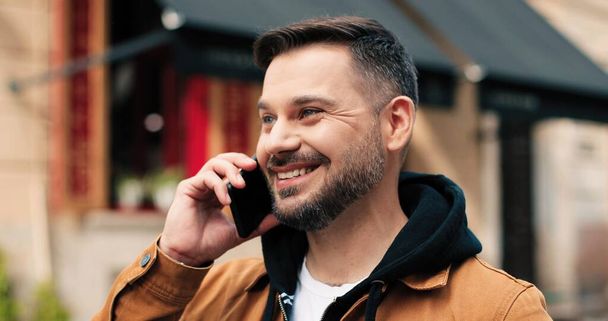 Portrait of young man talking smartphone outdoors. Beautiful caucasian guy using mobile phone while making call at the evening street with blurred background - Фото, зображення