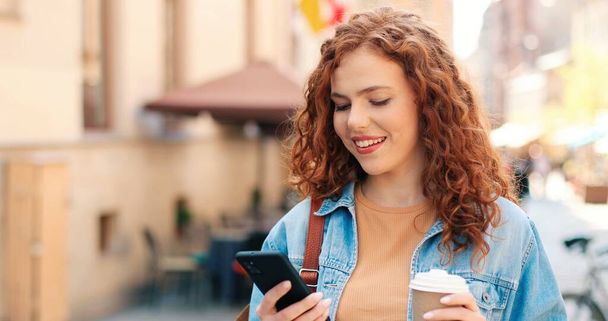 Retrato de caminar chica sonriente relajada usando ropa casual estilo de vida cómodo usando teléfono inteligente digital móvil y beber café al aire libre con fondo borroso - Foto, imagen