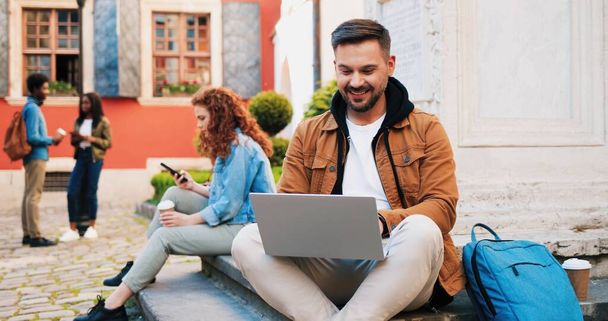 Jonge glimlachende brunette man dragen casual kleding met behulp van laptop computer buiten in de straat, terwijl zitten in de buurt van de muur en het verzenden van berichten. - Foto, afbeelding