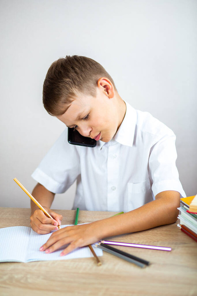 Το συναισθηματικό ξανθό αγόρι με το λευκό πουκάμισο μιλάει στο τηλέφωνο και γράφει κάτι σε ένα σημειωματάριο ενώ κάθεται στο γραφείο του. Τεχνολογία, επικοινωνία στο διαδίκτυο, ψυχαγωγικές δραστηριότητες για παιδιά  - Φωτογραφία, εικόνα