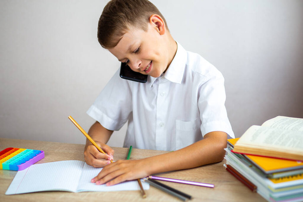 Emotional blond boy in a white shirt is talking on the phone and writing something in a notebook while sitting at his desk. Technology, internet communication, leisure activities for children  - Φωτογραφία, εικόνα